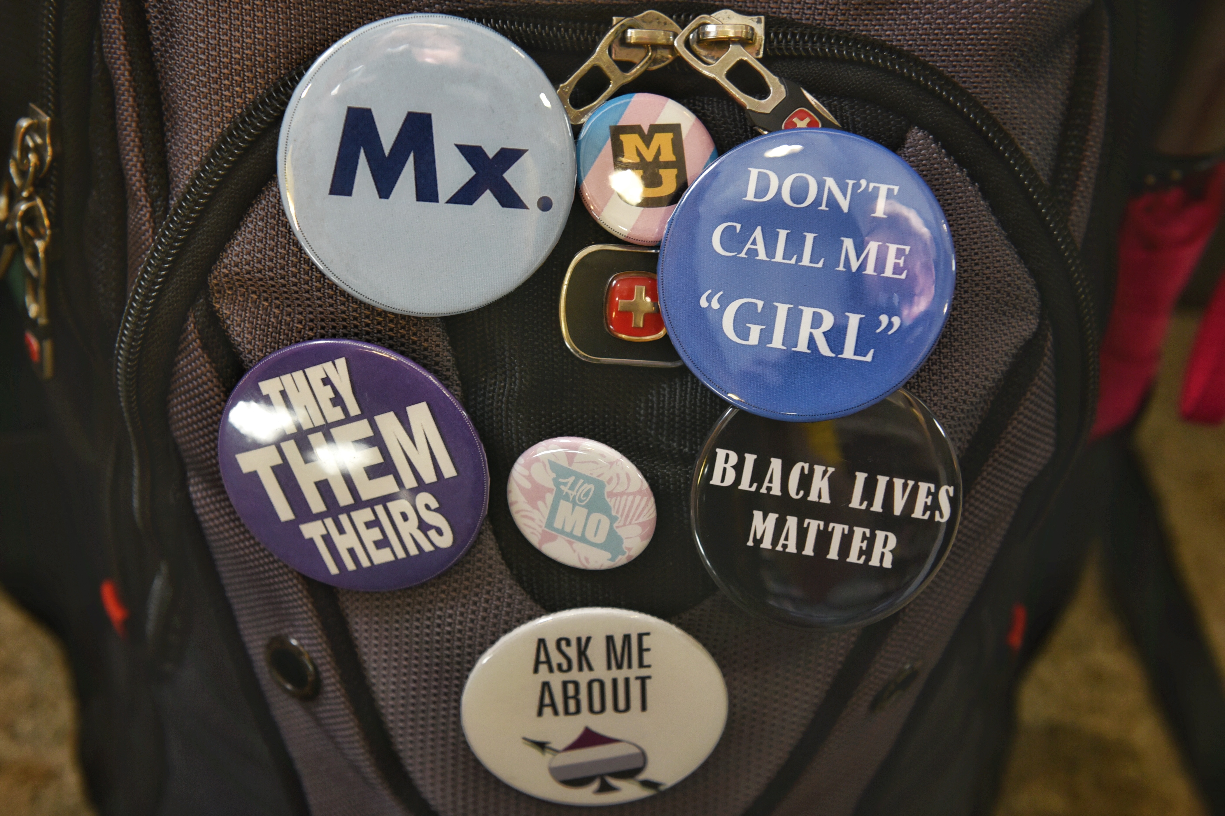 Buttons on Waldman's backpack, including "Mx. MU with a trans flag, Don't Call Me Girl, Black Lives Matter, They Them Theirs pronouns, and Ask Me About Aces and Aros"