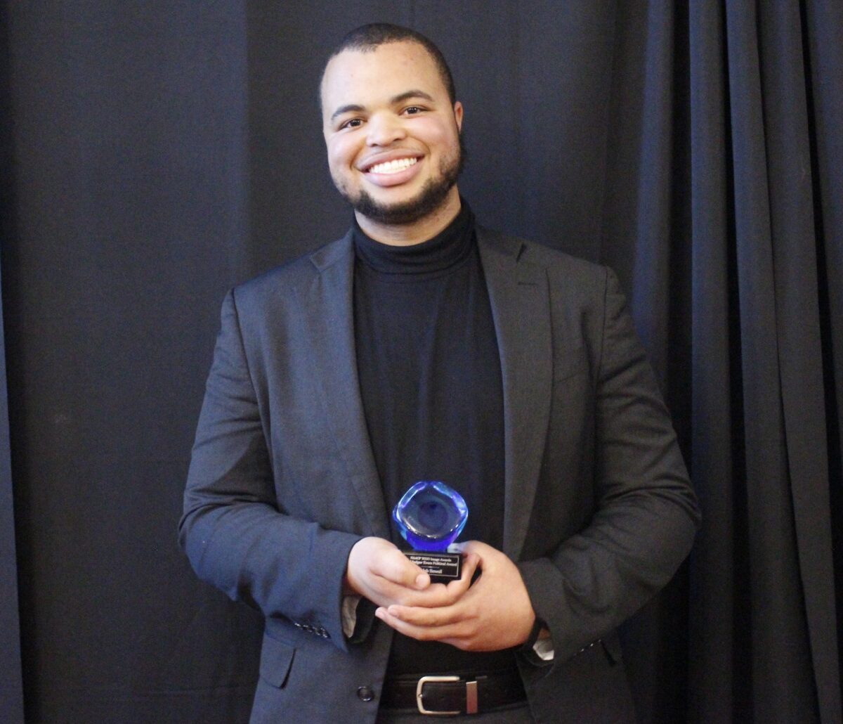 Caleb Sewell in a dark jacket and shirt holding the award.