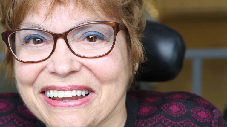 This is a photograph of Judy Heumann. She is sitting in a wheelchair, wearing glasses and facing directly at the camera. She is smiling.