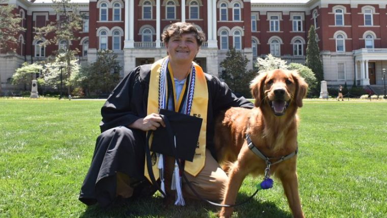 Colton Hicks in graduation regalia kneeling in front of Jesse Hall with his dog.